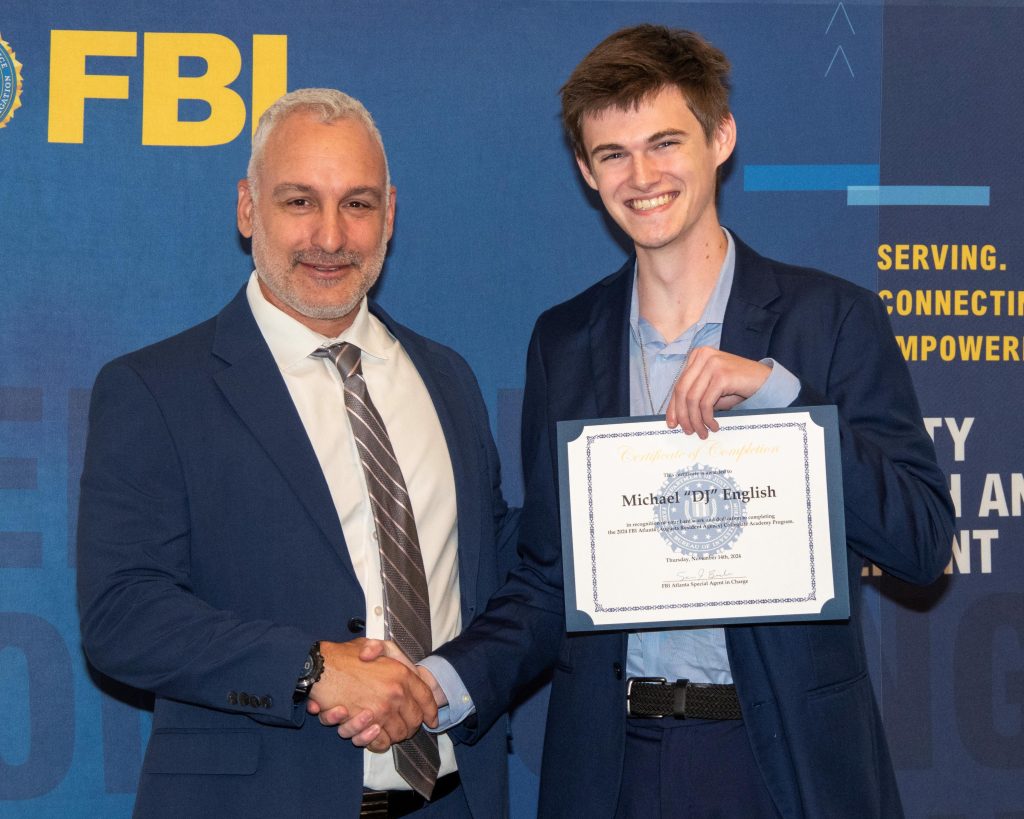 A man hands a college man a certificate of completion. There is a banner with the logo for the Federal Bureau of Investigation behind them.