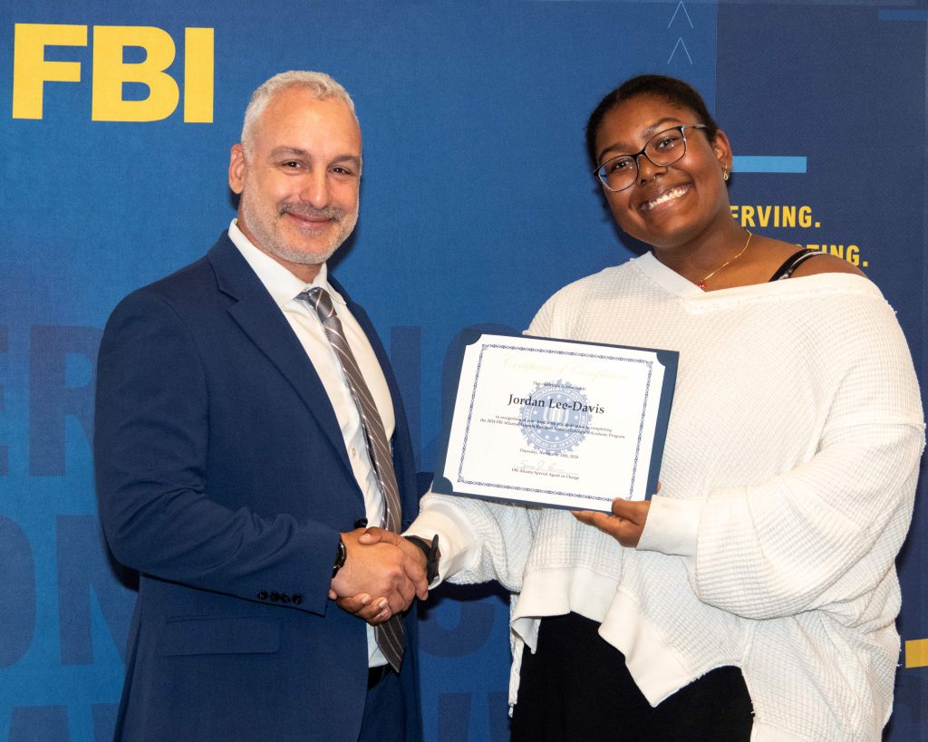 A man hands a college woman a certificate of completion. There is a banner with the logo for the Federal Bureau of Investigation behind them.