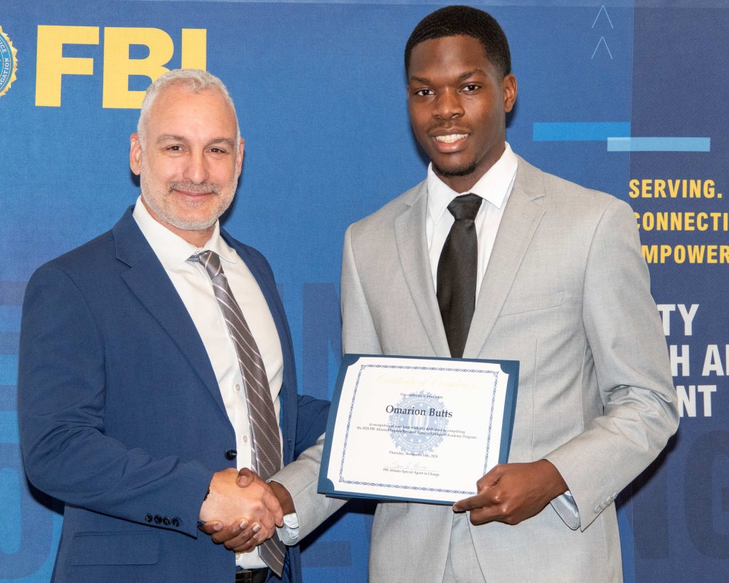 A man hands a college man a certificate of completion. There is a banner with the logo for the Federal Bureau of Investigation behind them.