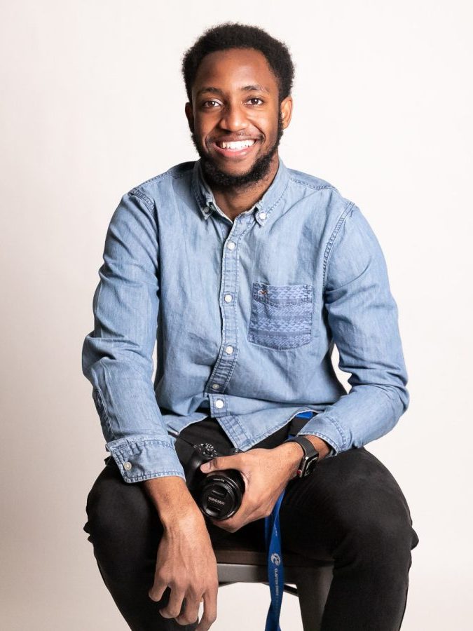 A man smiles for a photo while seated and holding a camera.