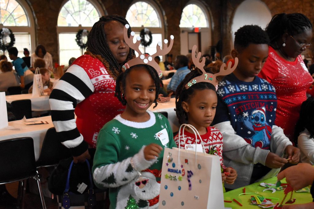 Three young children, two wearing paper reindeer antlers, work on arts and crafts at a table while their grandma watches.