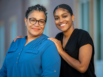 Two women standing outside a building. The younger woman has her hands on the shoulders of the other.