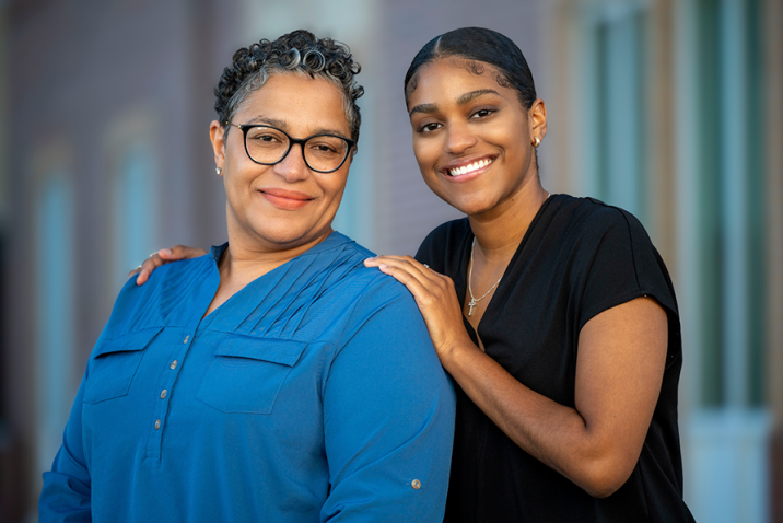 Two women standing outside a building. The younger woman has her hands on the shoulders of the other.