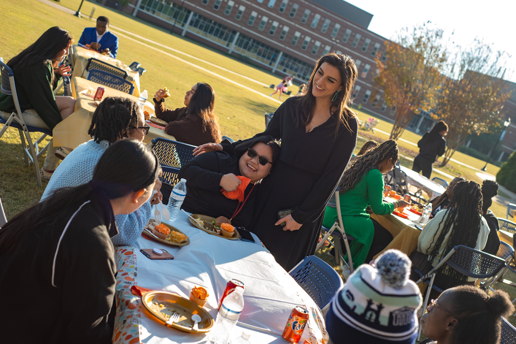 A woman stands next to an outside table where several college women are sitting and eating. One of the women is leaning in to hug the standing woman.