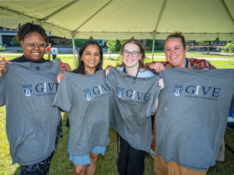 Four women stand under a large tent and hold up t-shirts.