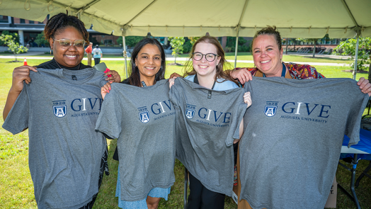 Four women stand under a large tent and hold up t-shirts.