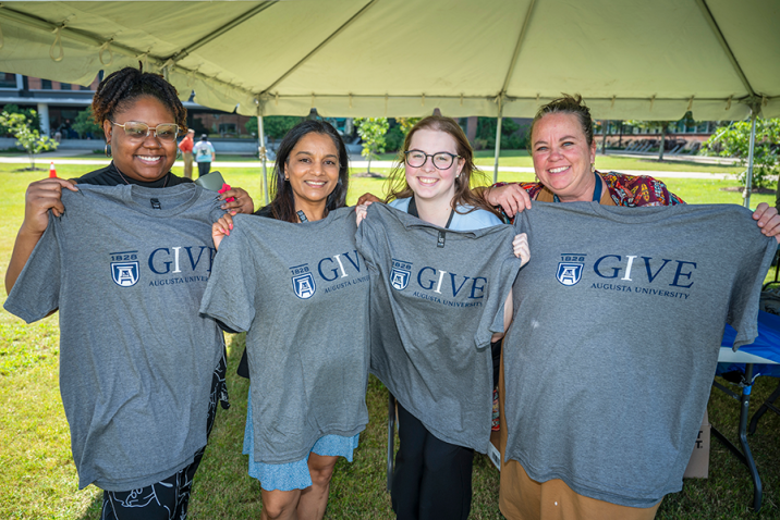 Four women stand under a large tent and hold up t-shirts.