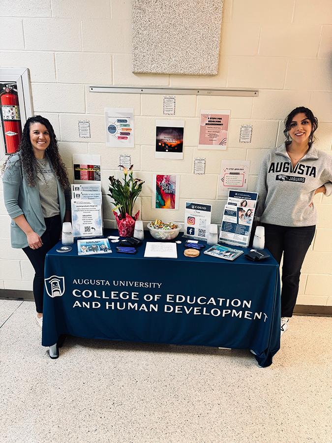 Two women wearing clothing branded for Augusta University stand on either side of a table. The table has information pamphlets about the College of Education and Human Development at Augusta University.