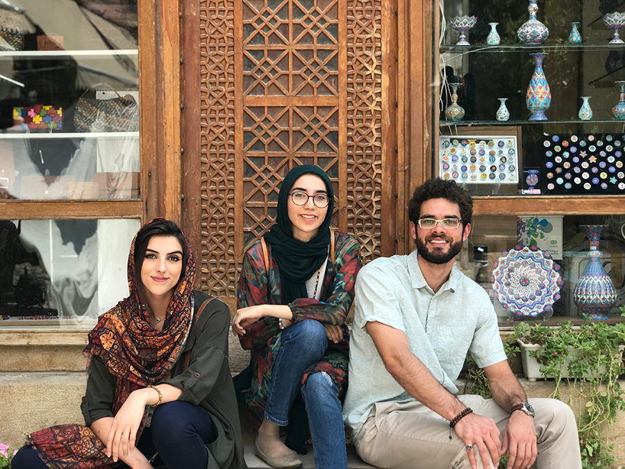 Two women and a man sit on a bench outside of a shop. There are colorfully-decorated vases and other trinkets in a window behind them.