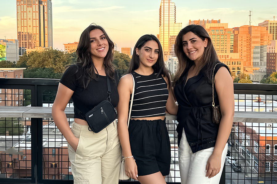 Three women stand outside on a patio with a city in the background.