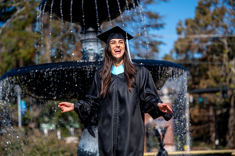 A college graduate wearing her graduation cap and gown stands outside in front of a large fountain while laughing.