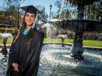 A college graduate wearing her graduation cap and gown stands outside in front of a large fountain.