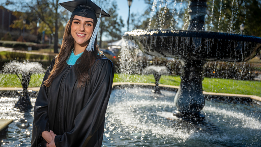 A college graduate wearing her graduation cap and gown stands outside in front of a large fountain.