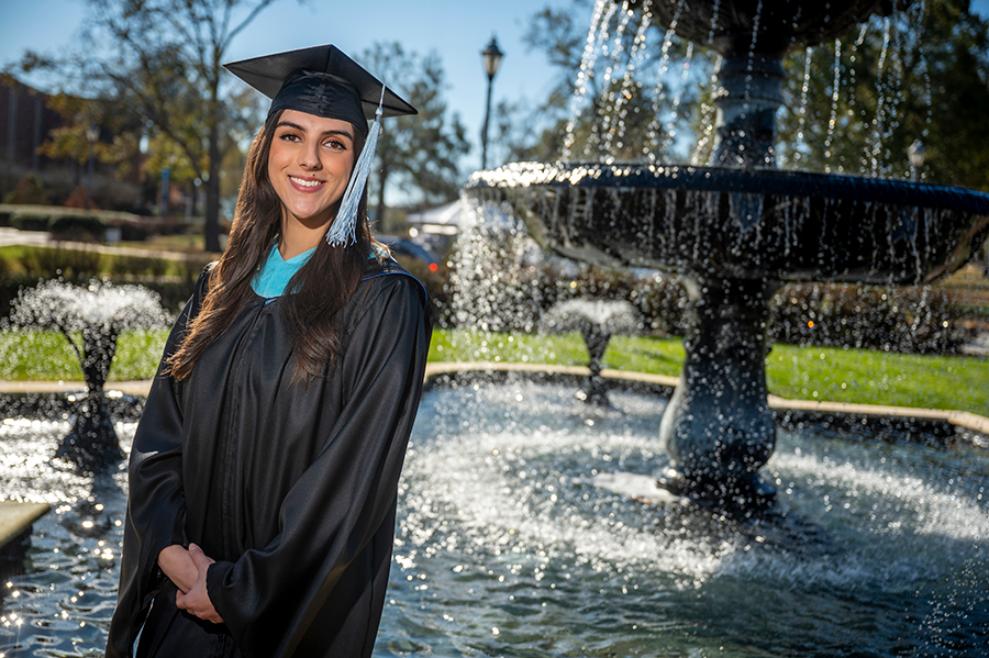 A college graduate wearing her graduation cap and gown stands outside in front of a large fountain.