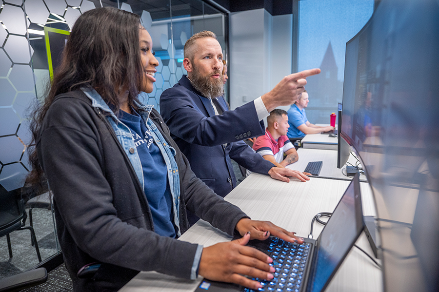 A man and a woman stand at a tall table with a computer on it. The man is pointing to an extra screen while the woman types on the computer's keyboard.