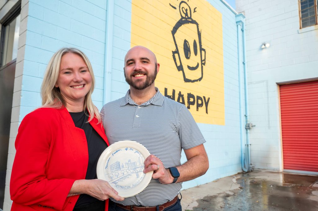 A woman and a man hold both sides of a painted plate in front of a mural. 