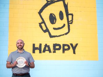 A man holds a painted plate in front of a mural.