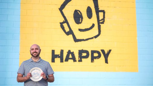 A man holds a painted plate in front of a mural.