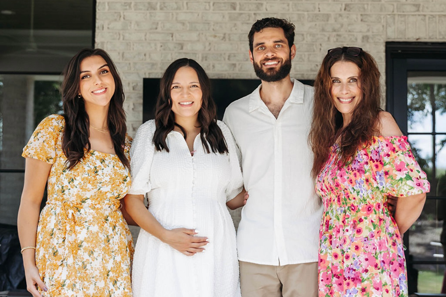 Three women and one man stand outside on a patio. One of the women is pregnant and holding a hand gently over her belly.