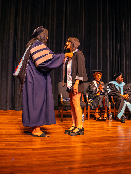 Two women stand on a stage. One woman is dressed in full college graduation regalia signaling she holds a doctorate, and she is placing a stole around the shoulders of the other woman.