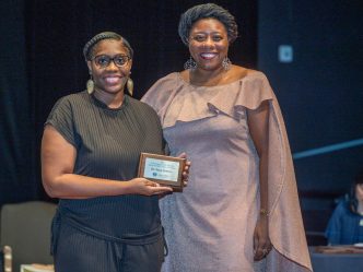 two women smile at the camera while the woman on the left holds an award