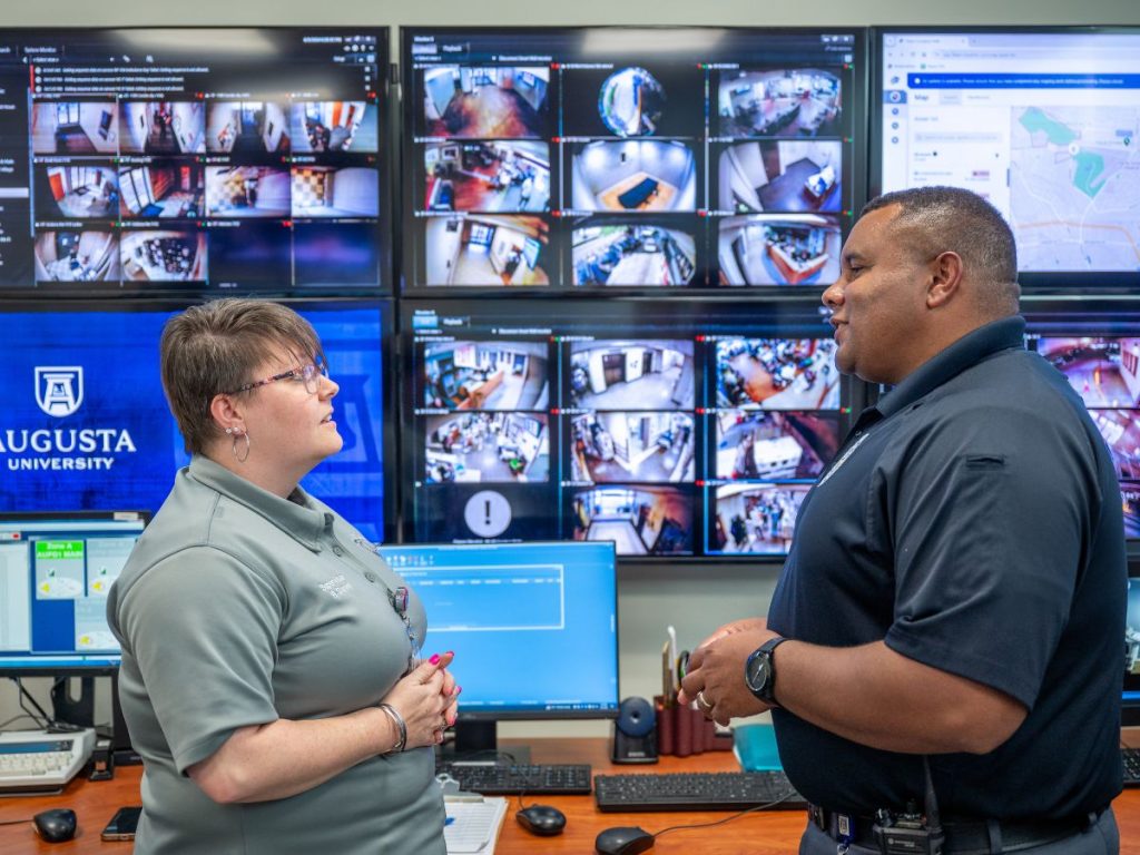 Two police officers stand together talking in front of a wall of monitors.