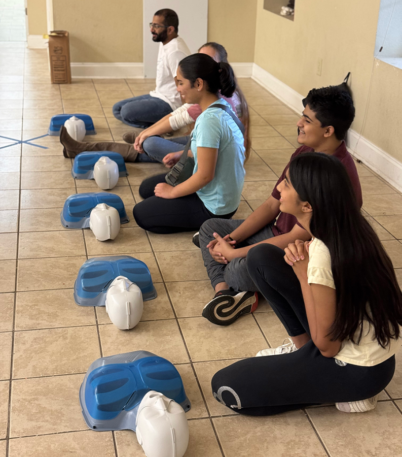 Young boys and girls sitting on the ground in a line. There are medical mannequins in a line in front of each of them.