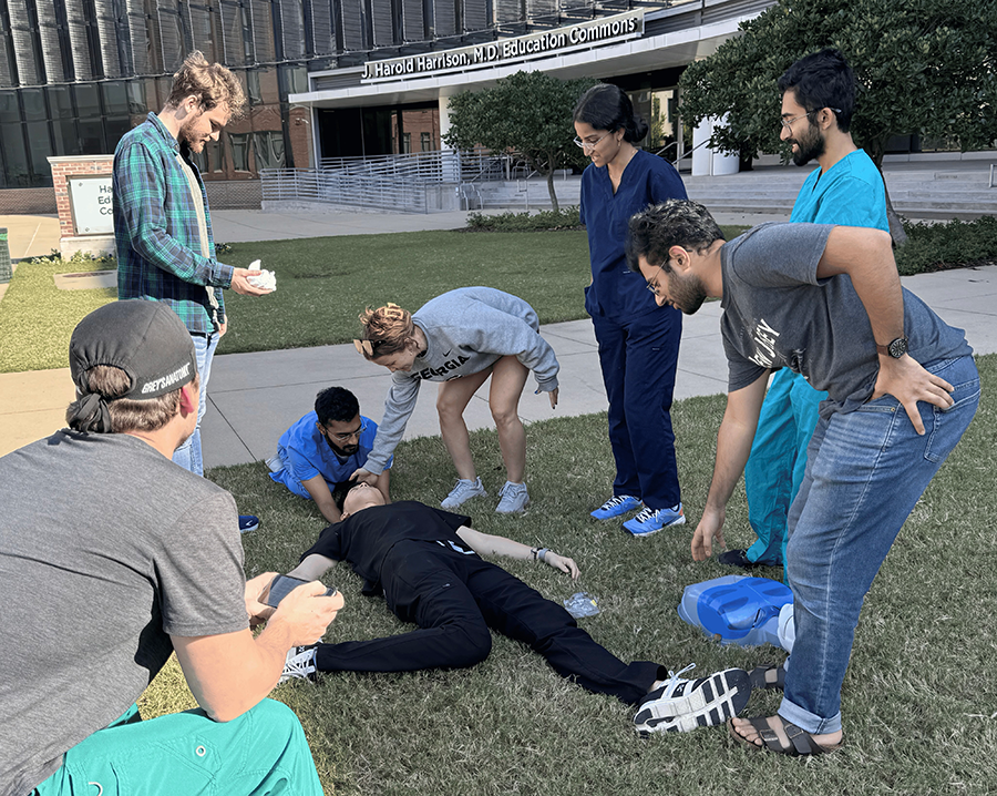 A group of seven medical students gather in a courtyard. A 
mannequin is laying on the ground, injured. The students are tending to its injuries.