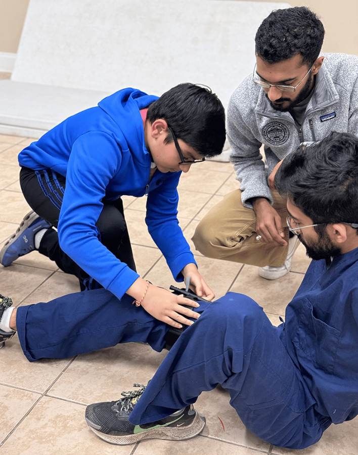 A young boy applies a medical tourniquet on a man's leg. Another man watches over them and observes.