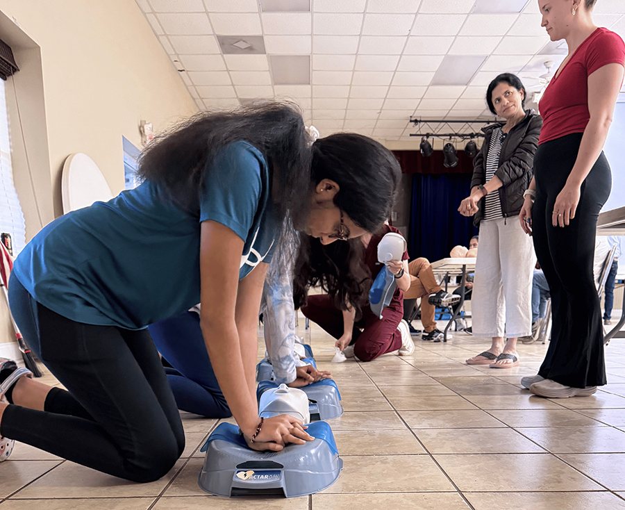 A line of school aged children practice CPR on mannequins. An instructor oversees them.