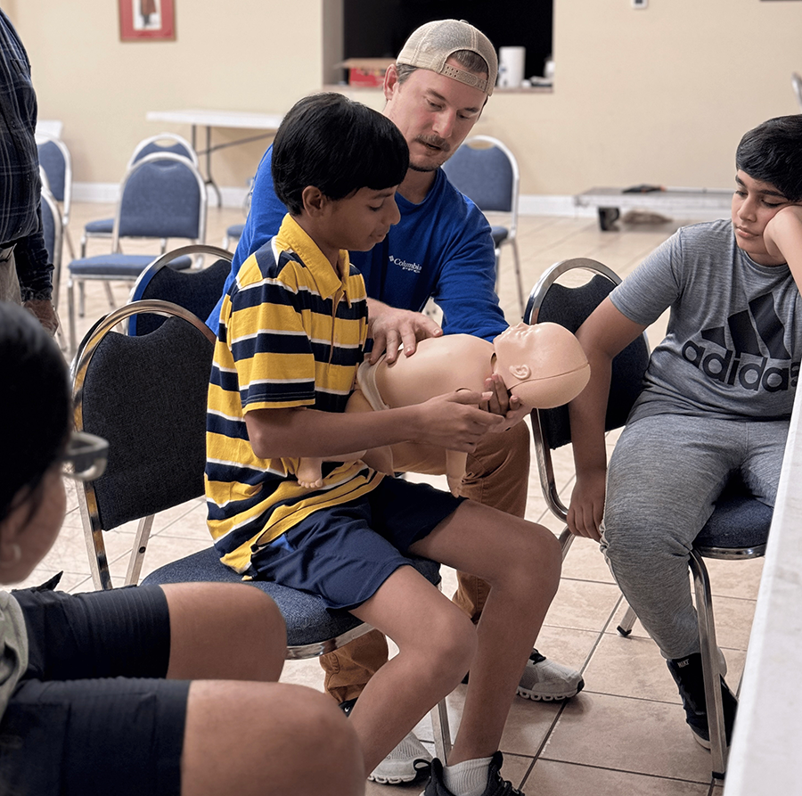 A grade school student holding a mannequin infant. An instructor shows him how to handle it properly.