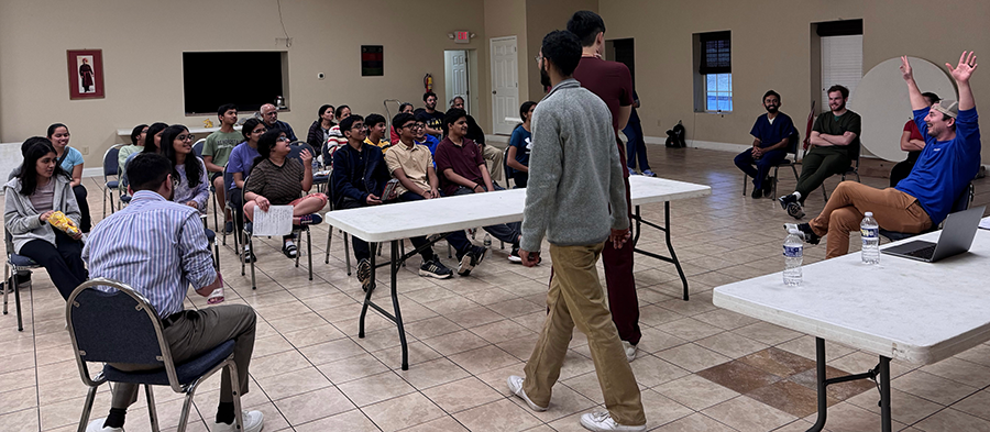 A group of about 25 grade school students sit and watch medical instructors teach first aid lessons.