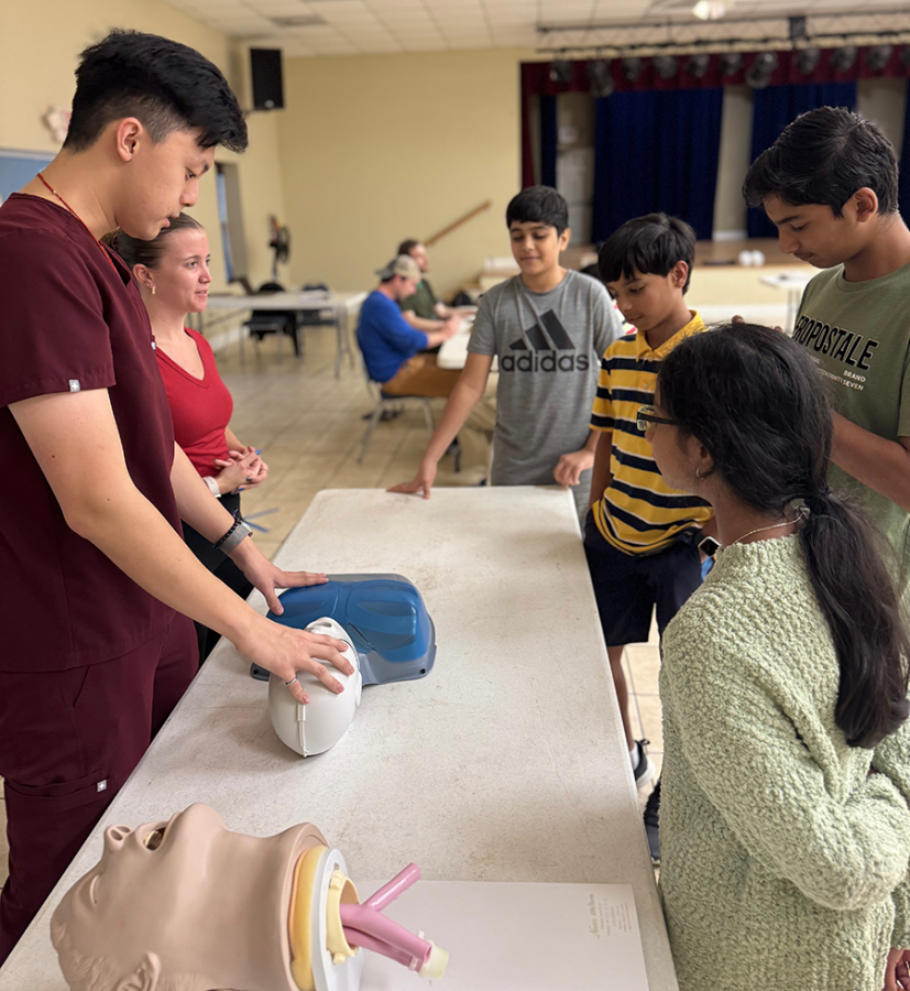 Four grade school students gather around a table, where two medical students are showing them how to do CPR on a mannequin.