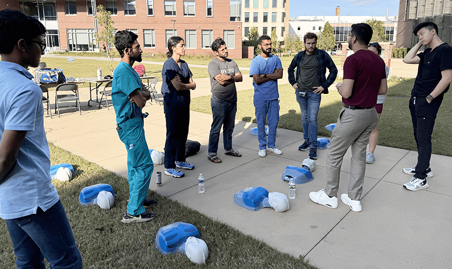 A group of nine medical students listening to an instructor in a courtyard. There are medical mannequins on the ground.