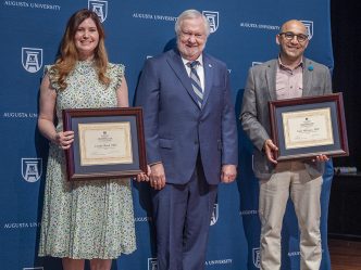 A woman and a man stand in front of a navy blue Augusta Univeristy backdrop while each holding a framed certificate as a man in a suit stands between them, all smiling at the camera.