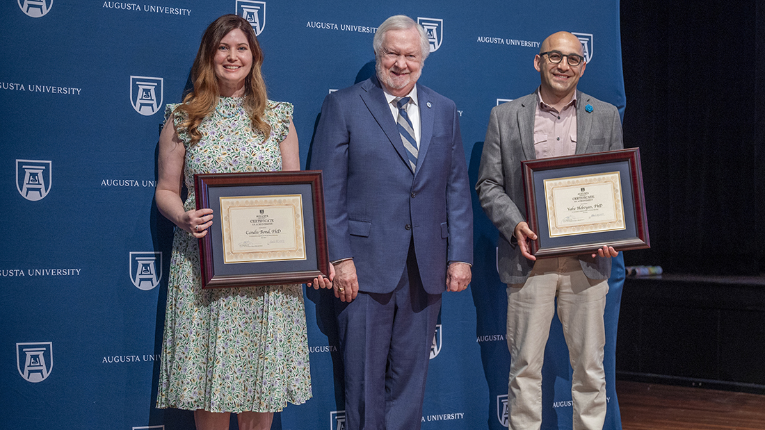 A woman and a man stand in front of a navy blue Augusta Univeristy backdrop while each holding a framed certificate as a man in a suit stands between them, all smiling at the camera.