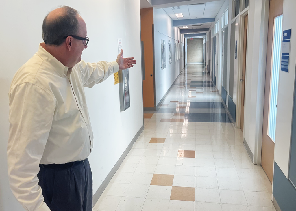 A professionally dressed man points at a series of doors down a hallway.