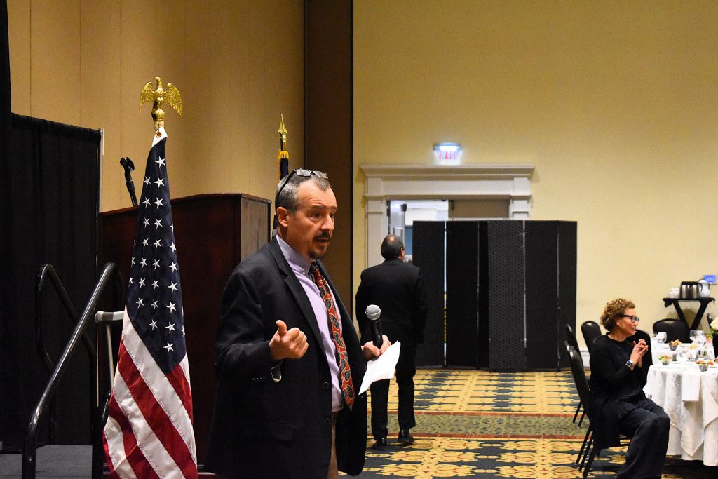 A man wearing a suit stands in front of a large group of people and speaks into a microphone.