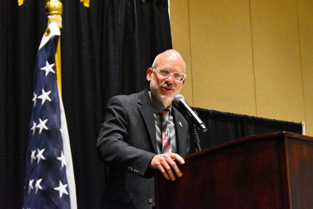 Man in a suit stands at a podium with a microphone and delivers remarks to a large group of people.