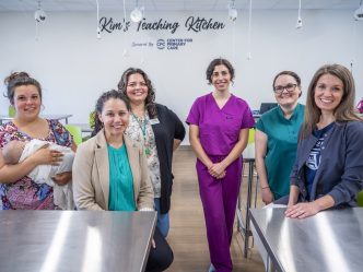 Six women, one holding a baby, stand in a large industrial kitchen