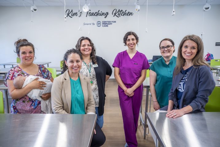 Six women, one holding a baby, stand in a large industrial kitchen