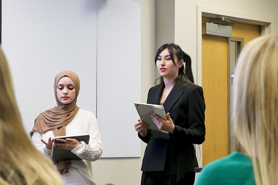 Two female college students stand at the front of a classroom and present on a project they completed. Both are holding electronic tablets.