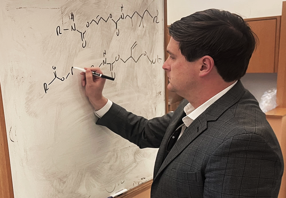 A man in a suit writes a scientific formula on a dry erase board.