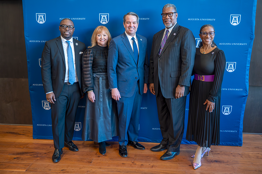 Three men wearing suits and two ladies stand in front of a backdrop with the logo for Augusta University.