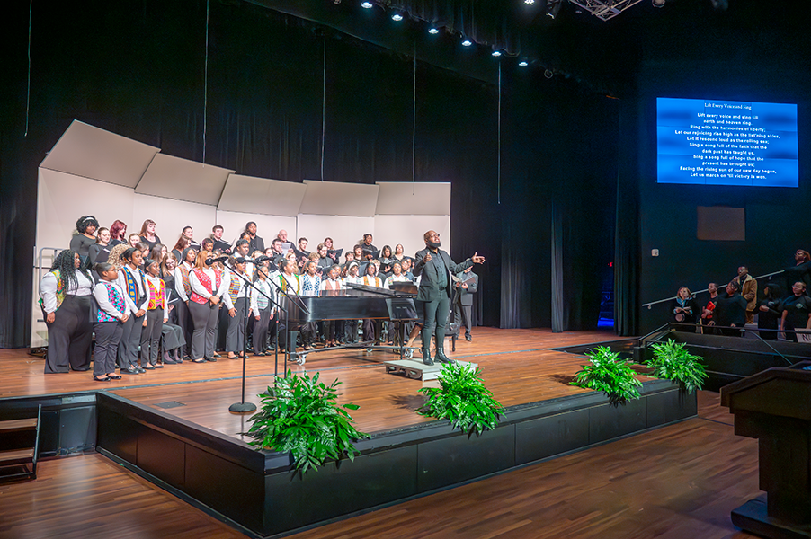 A large choir of young people stands ready to sing as their director faces the crowd to invite them to join in song.
