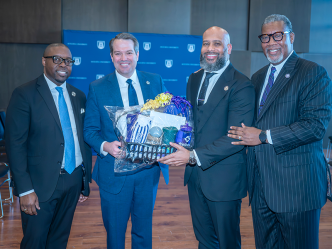 Four men in suits stand together with two of them holding a gift basket.