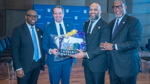 Four men in suits stand together with two of them holding a gift basket.