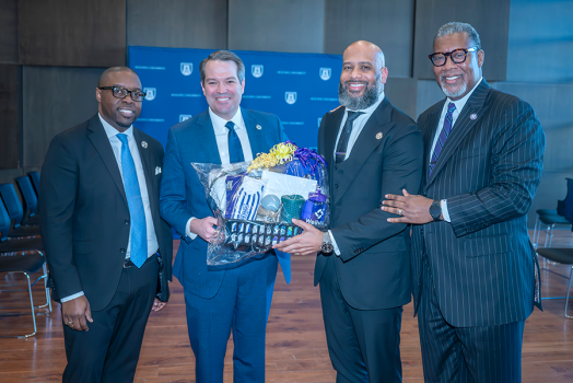 Four men in suits stand together with two of them holding a gift basket.