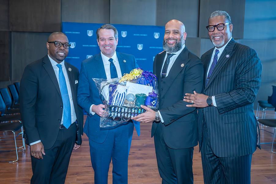 Four men in suits stand together with two of them holding a gift basket.