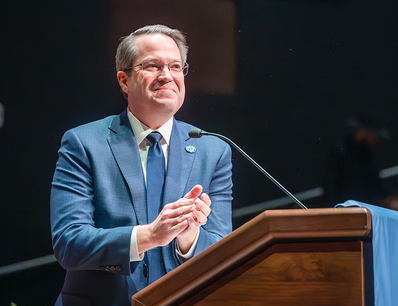 A man in a suit behind a lectern claps his hands.